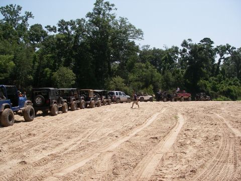 1995 Ford Bronco - A group of off-road vehicles are lined up on a sandy terrain, with trees forming the backdrop.