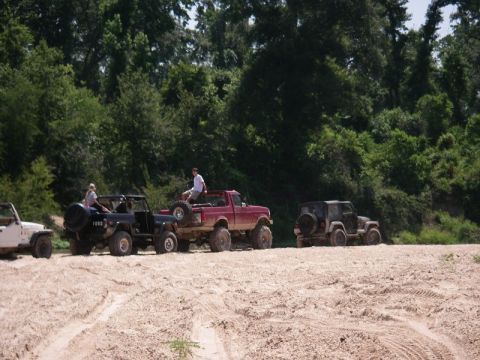 1995 Ford Bronco - A group of off-road vehicles are gathered on a sandy track, with lush green trees forming the backdrop.