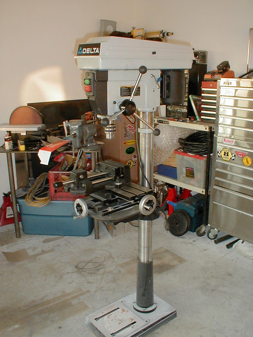 Delta drill press with a milling vise attached, set up in a garage with tool chests and other equipment