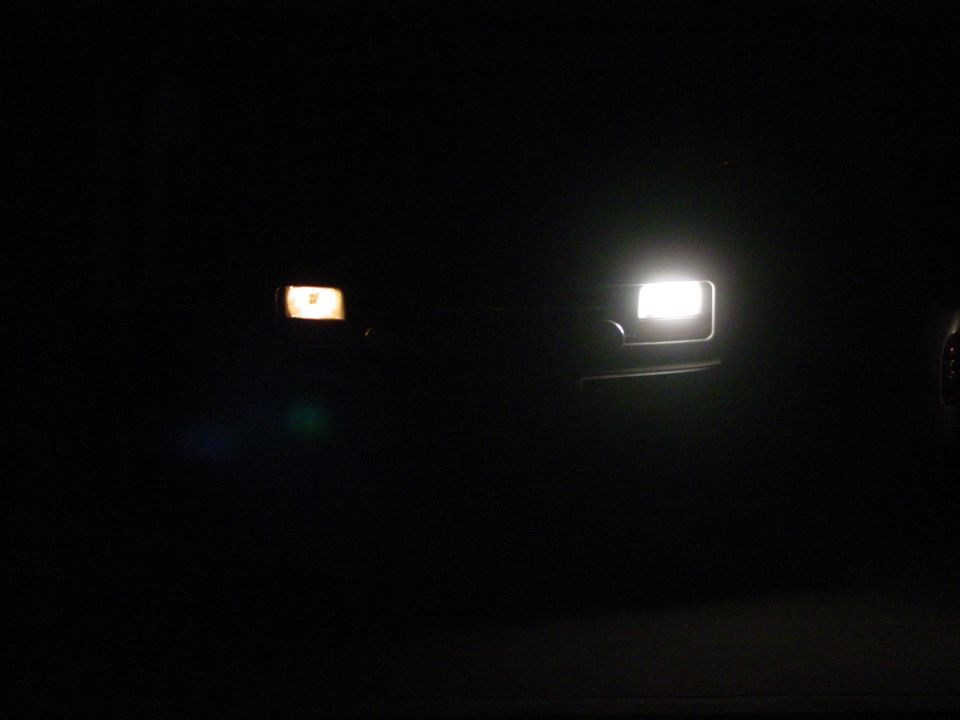 Two vehicle headlights illuminated at night. One is bright white, the other is a dimmer amber