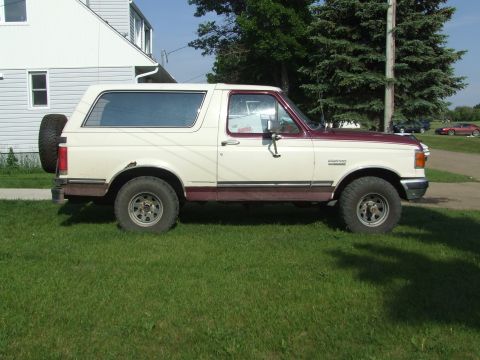 1990 Ford Bronco - Side profile of a two-tone 1990 Ford Bronco, parked on a grassy lawn next to a house.