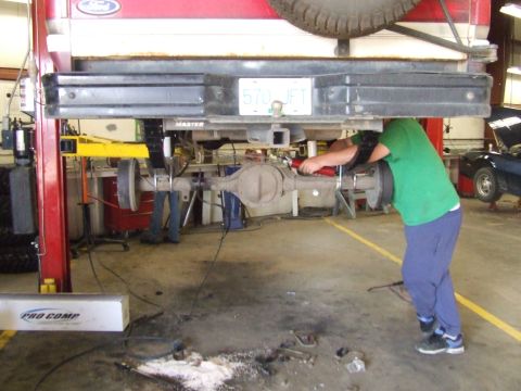 1990 Ford Bronco - Mechanic works on the rear axle of a vehicle suspended on a lift in a garage setting.