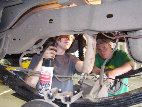 1990 Ford Bronco - Two mechanics work on the underside of a vehicle, one applying lubricant to the suspension system.