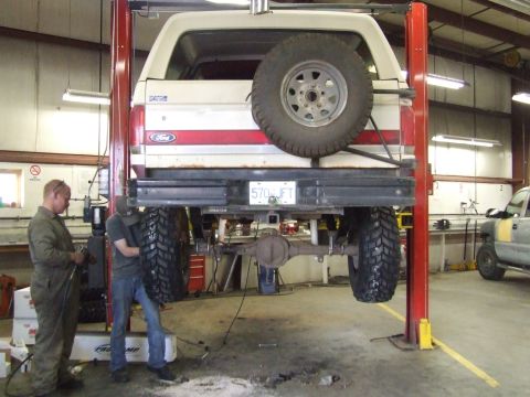 1990 Ford Bronco - Mechanics work on the undercarriage of a lifted Ford Bronco in a garage setting.