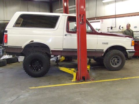 1990 Ford Bronco - A two-tone Ford Bronco is raised on a hydraulic lift in a garage, revealing its undercarriage and large tires.