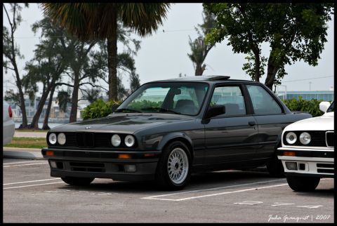 1989 BMW 3 Series - A dark gray coupe is parked in a lot next to another vehicle, with trees and buildings in the background.