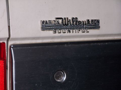 1990 Ford Bronco - Close-up of a chrome dealer badge reading "MARION WILLEY & SON, BOUNTIFUL" screwed to a white body panel above a brushed stainless trim pie…