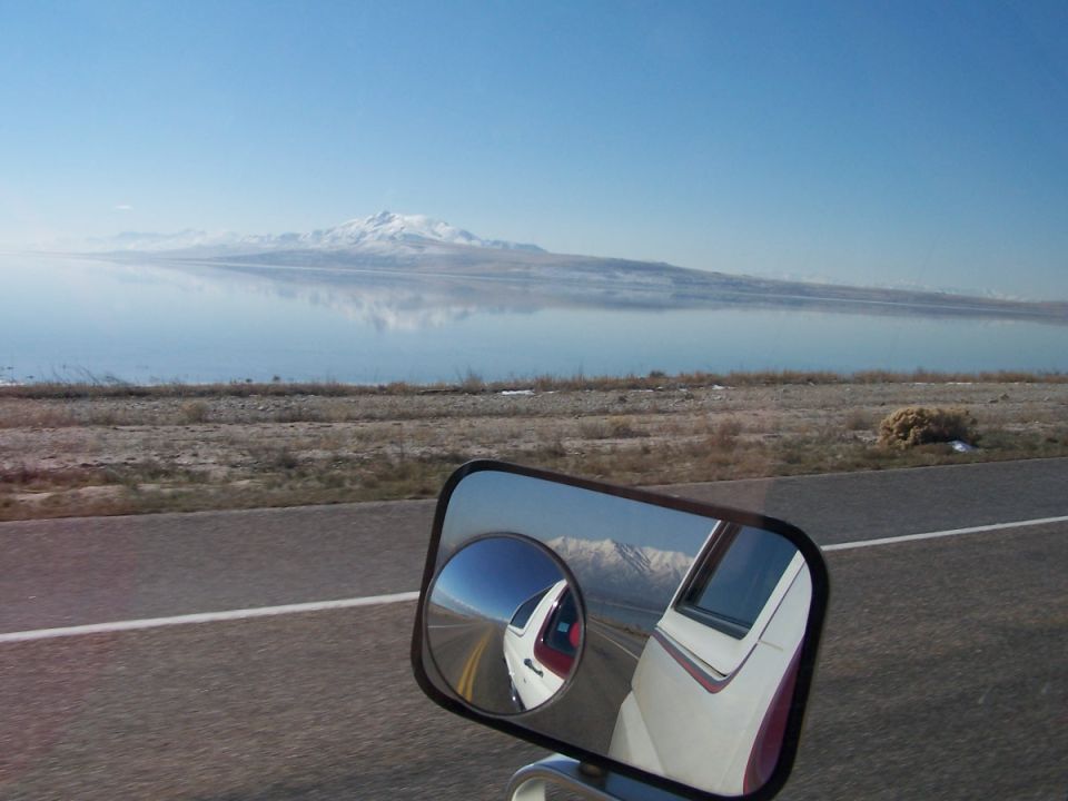 Towing mirror with convex spot mirror reflecting a white trailer beside a salt lake and snowy mountains