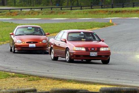 1997 Ford Taurus - Two red Ford Taurus sedans running side by side through a corner at a road course, both with helmeted drivers and numbered windshield decal…