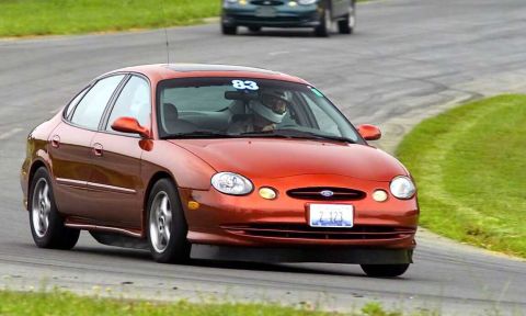 1997 Ford Taurus - A red-orange oval-body Ford Taurus sedan wearing number 83 on the windshield corners through a track-day turn, the helmeted driver visible…