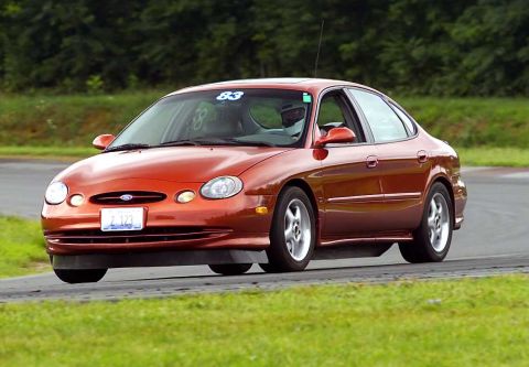 1997 Ford Taurus - Orange-red Ford Taurus sedan running a track session, caught leaning through a corner on a paved course lined with grass and trees. Blue '8…