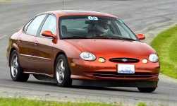 1997 Ford Taurus - Small, low-resolution three-quarter front shot of a copper/orange Ford Taurus sedan on a paved course with a grassy verge behind, showing a…