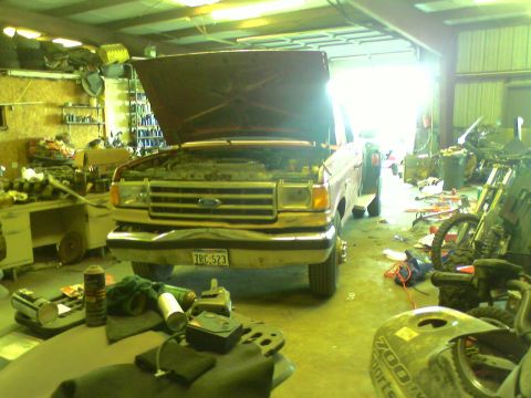 1982 Ford Bronco - A red pickup truck sits in a garage with its hood open, showing the engine bay. Other vehicles and tools are visible in the cluttered space.