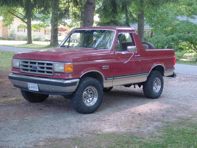 Red 1987 Ford Bronco with tan lower body trim, lifted suspension, and aftermarket wheels parked on gravel