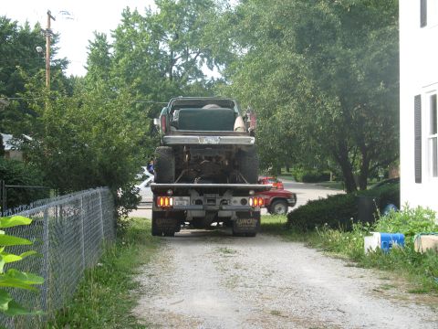 1978 Ford Bronco - A flatbed truck transports a lifted SUV down a gravel driveway, with a red pickup truck in the background.