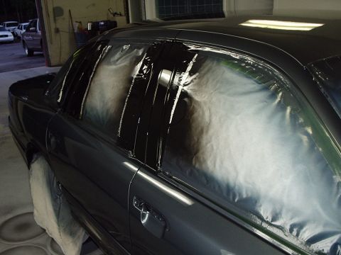 1996 Ford Crown Victoria - A dark sedan's side windows are taped off and covered with plastic sheeting during a refinishing process.