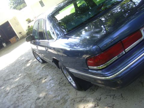 1996 Ford Crown Victoria - The rear of a dark blue Ford Crown Victoria is visible, parked on a dirt surface with a garage in the background.