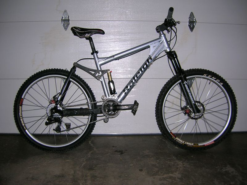 Silver Raleigh full-suspension mountain bike with black fork and tires, parked in front of a garage door