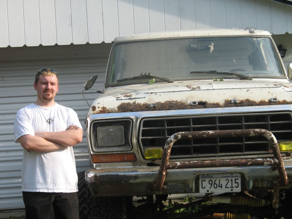 Guy in white t-shirt arms crossed next to a beat-up white Ford pickup. Truck has rust on hood and grille, yellow fog lights, and a brush guard