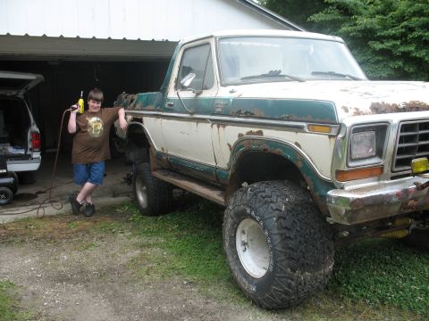 1978 Ford Bronco - A boy stands with a drill beside a heavily modified 1978 Ford Bronco, showing off its lifted suspension and off-road tires.