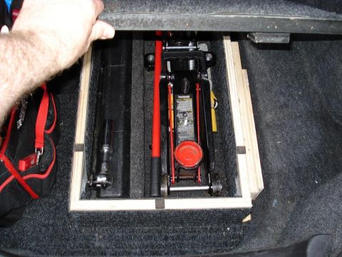 1999 Ford Crown Victoria - A hand lifts a carpeted trunk-floor lid to reveal a custom wood-framed storage well holding a red-and-black low-profile floor jack, a break…