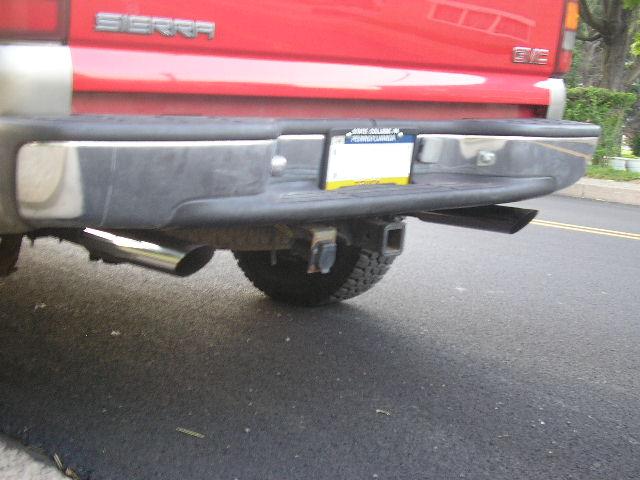Red GMC Sierra pickup truck rear bumper with dual exhaust pipes and a trailer hitch. License plate visible