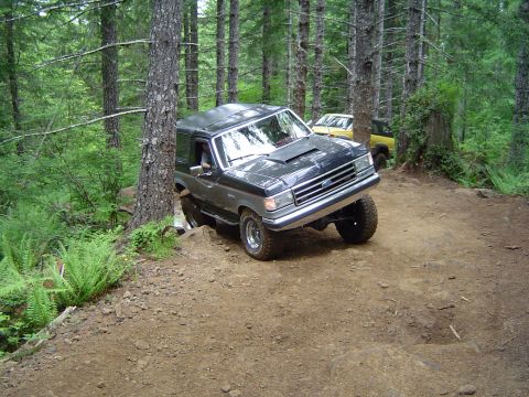 1989 Ford Bronco - A dark gray Ford Bronco navigates a challenging dirt trail in a forest setting, with another vehicle visible in the background.