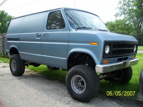 1978 Ford E250 - Three-quarter front view of a light blue lifted 4x4 Ford Econoline van parked at the edge of a driveway, sitting high on all-terrain tires…