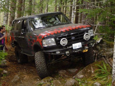 1989 Ford Bronco - 1989 Ford Bronco navigating a wooded trail with large tires and custom front bumper.