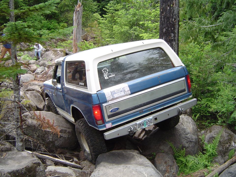Blue and white Ford Bronco crawling over boulders on a forest trail, rear three-quarter view