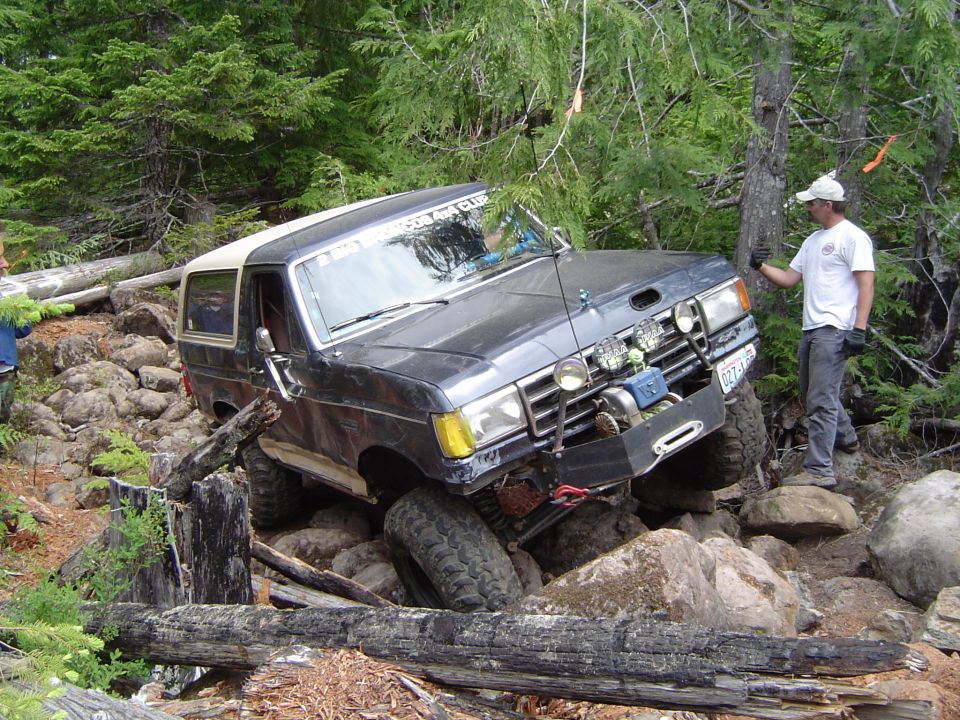 Dark Ford Bronco with winch crawling over rocks on a wooded off-road trail, man spotting.