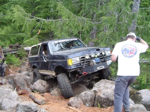1989 Ford Bronco - A modified Ford Bronco tackles a rocky trail, with a man in a Bronco Club shirt observing.