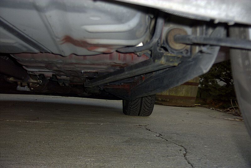 Underside view of a vehicle showing rear suspension, frame and a tire, photographed from beneath on concrete