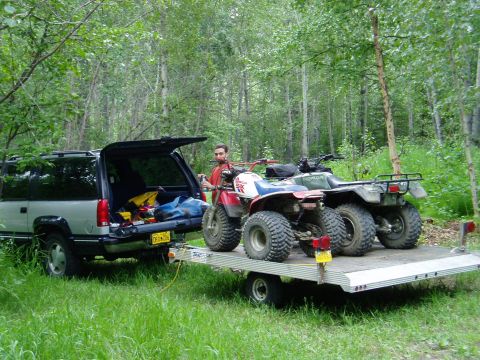 2000 Ford E350 - A man stands by a trailer loaded with ATVs, hitched to a silver SUV in a forest clearing.
