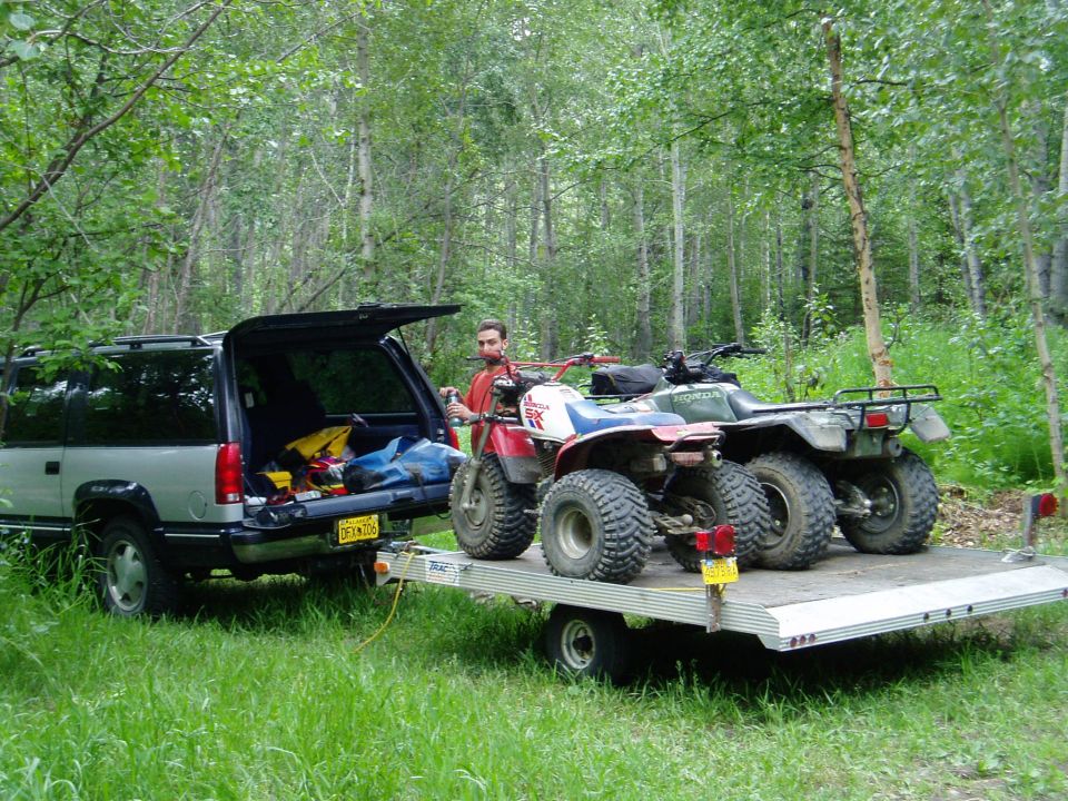 Silver SUV with open tailgate parked next to a trailer carrying two ATVs in a wooded area