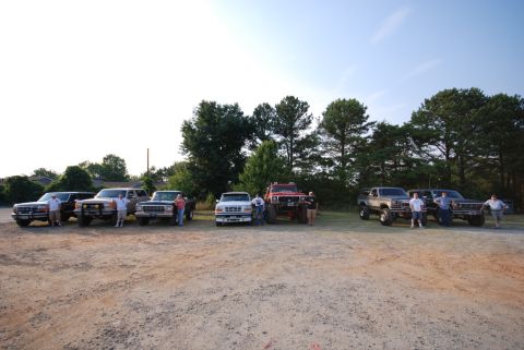 1996 Ford Bronco - A group of trucks and SUVs are gathered together in a dirt lot with trees in the background.
