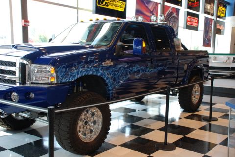 1996 Ford Bronco - A lifted blue pickup truck with custom flame graphics is displayed indoors on a checkered floor.