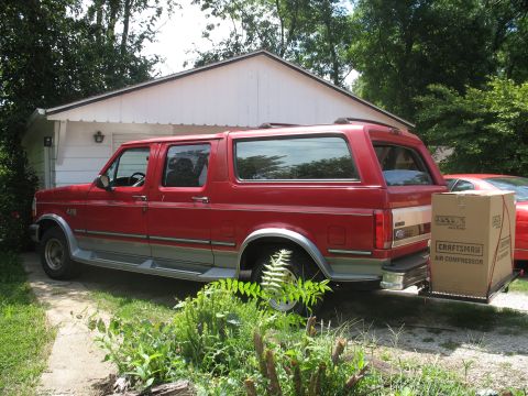 1994 Ford C-150 (Centurion Conversions) - A red SUV is parked in front of a white garage, with a large box on its hitch.