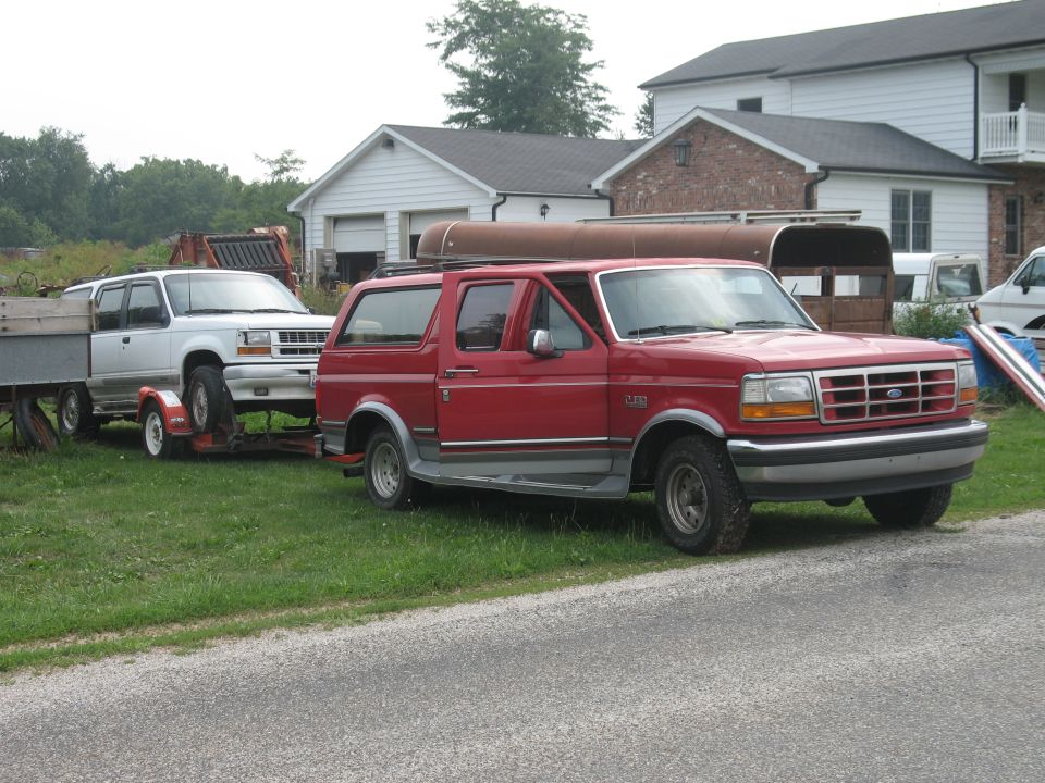 Red Ford Bronco with a canoe on top, towing a white Ford Explorer on a trailer