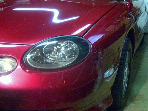 1997 Ford Taurus - Close-up of the driver-side oval headlamp and amber corner marker on a dark red sedan, shot low from the front corner in a garage. The clea…