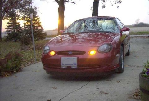 1997 Ford Taurus - Front three-quarter view of a dark red four-door sedan parked on a concrete driveway at dusk, with a pair of amber round fog lamps lit low…