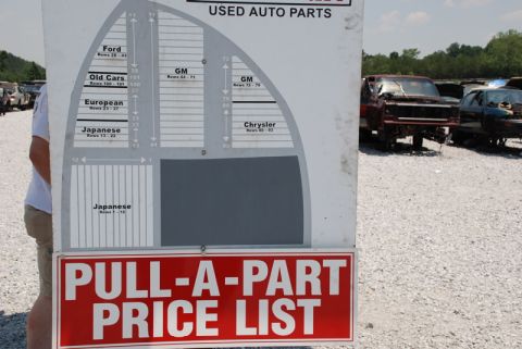 1996 Ford Bronco - A sign at a junkyard lists prices and sections for used auto parts, with several vehicles in the background.