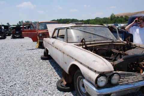 1996 Ford Bronco - A weathered 1960s sedan sits in a junkyard, its hood ajar and wheels removed.