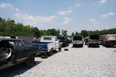 1996 Ford Bronco - A variety of vehicles are parked in a junkyard under a partly cloudy sky.