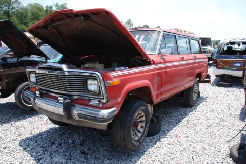 1996 Ford Bronco - A red Jeep Grand Wagoneer sits in a junkyard with its hood open, surrounded by other vehicles.