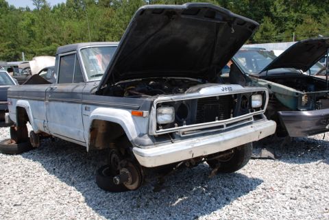1996 Ford Bronco - A weathered Jeep pickup truck sits in a junkyard with its hood open and missing wheels.
