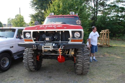 1996 Ford Bronco - A heavily modified red Ford truck with oversized tires and a humorous bumper sticker is parked next to another vehicle.