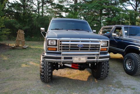1996 Ford Bronco - A lifted Ford Bronco is parked next to another SUV, with a wooden structure in the background.