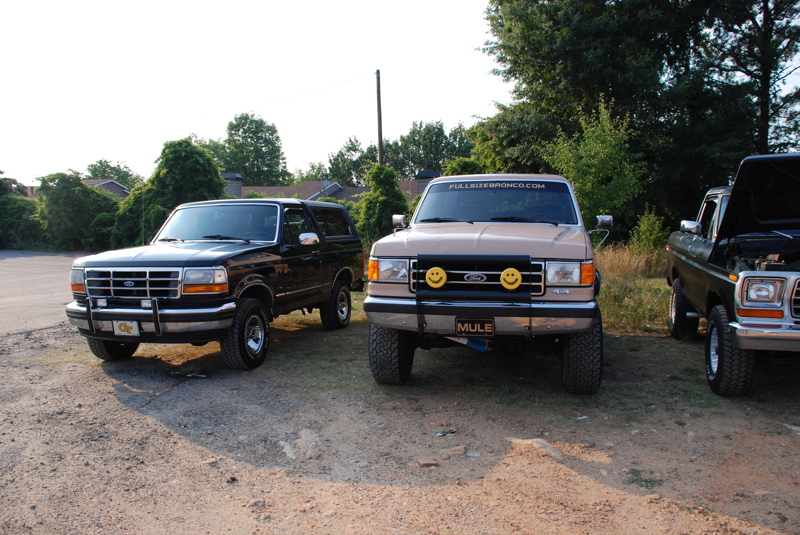 Three Ford Broncos parked side-by-side. The leftmost is black, the middle is tan with smiley face lights, and the rightmost is dark green with its hood open