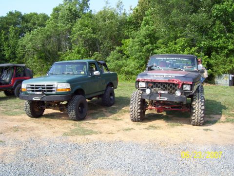 1978 Ford Bronco - Two modified SUVs are parked side-by-side on a dirt lot with trees in the background.